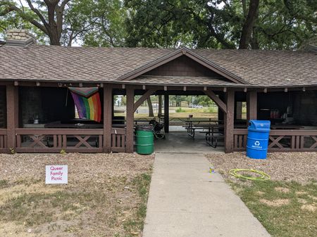 A covered picnic shelter in a park with a sign in front reading 'Queer Family Potluck Picnic' and a progress pride flag hanging from a rafter