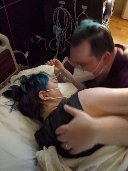 a person with short, dyed blue hair with dark roots wearing a face mask lays in a hospital bed. Another person wearing a mask is nestled close to comfort the person laying down