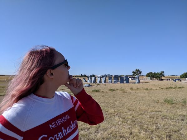Cassey, who appears in the photo as a person with long red hair an sunglasses, looks towards Carhenge in the distance, a replica of stonehenge made entirely of old cars painted grey