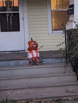 on the concrete steps to a front porch, a figure has been created from a pumpkin, half eaten with a painted on scared face, a white upturned plastic tub, and some crumpled up pants