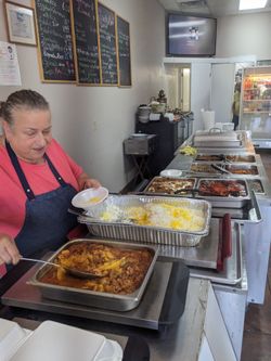 an older woman with greying dark hair pulled back into a bun serves meat and veggies in thick colorful sauces  from big roasting pans