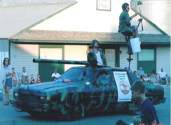 an old car has been painted in camo colors, even the windows. A guy in a camo jacket and helmet is sticking out the sunroof, and there's a fake cannon over the hood. The hot dog slingshot is mounted over the trunk with a camo-clad rider about the launch a hot dog