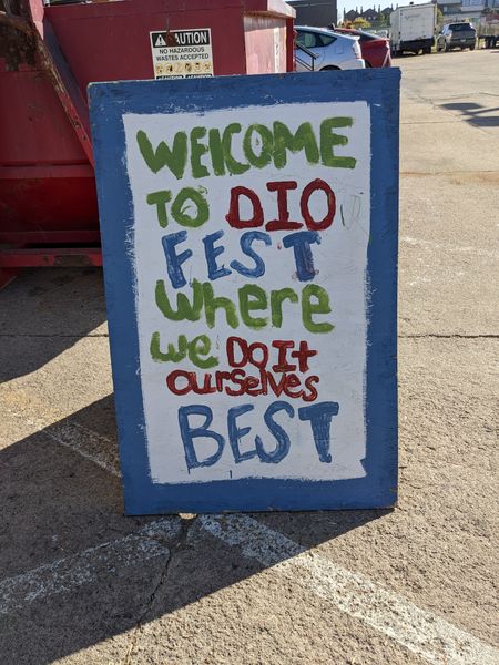 a hand-painted sandwich board sign in alternating primary colors reads 'Welcome to DIO Fest where we Do It Ourselves best'