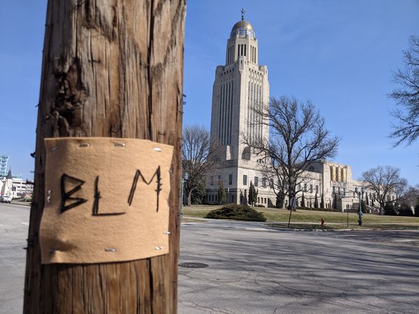 A piece of felt with BLM stitched on it is stapled to a pole in the foreground, with the Nebraska state capitol building rising in the background