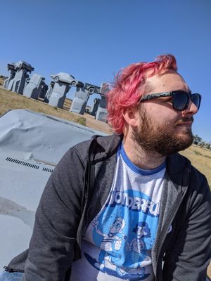 a person with a beard and short pink hair, wearing sunglasses, looks away from the camera. they're sitting on a grey-painted car, and Carhenge appears in the background