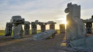 a large replica of Stonehenge made of grey-pained vintage cars against a sunset