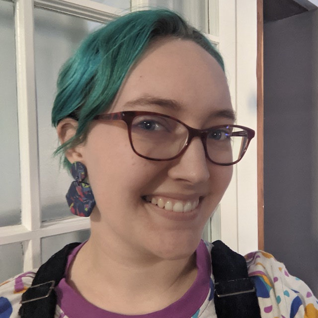 Cassey, a person with short blue-green hair and red glasses, smiles and shows off blue and pink two-tiered hexagonal earrings.