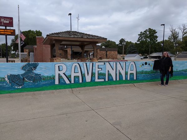 Cassey stands in front of a mid-chest-height mural of the word 'Ravenna' painted over a river background
