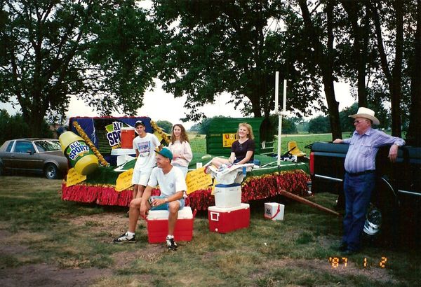 a float on a trailer where it's like a football field but the ground is wavy, and there are big bottles of a drink called All Sport. Several 90s people are standing around. The date on the photo says 87 as the year.
