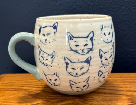 A cream colored mug with blue cat face line drawings printed all over the body. The handle is a pale blue.
