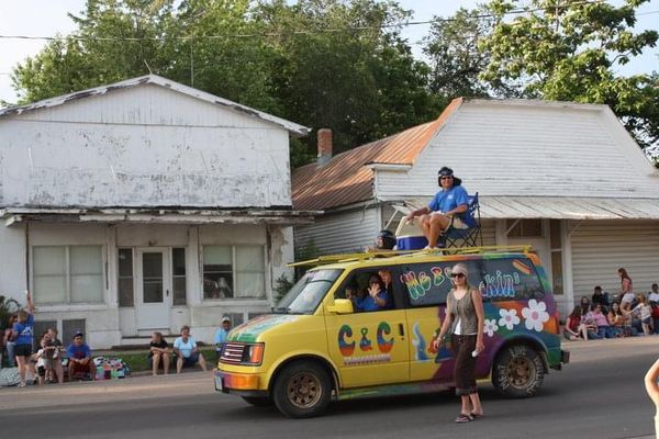 A van painted in floral hippie colors has a guy sitting in a lawn chair on top of it. The guy is reaching into a cooler. A blonde teenager in a hippie outfit walks alongside the van.
