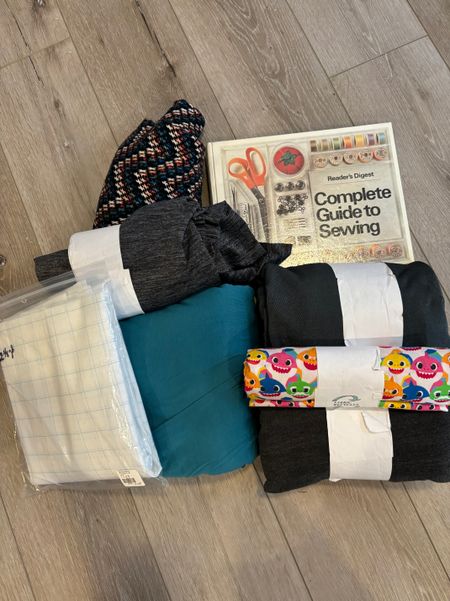 a pile of stuff including some knit fabric in various colors and a roll of baby shark patterned cotton, some swedish tracing paper in a plastic bag, and the Readers Digest Complete Guide to Sewing book