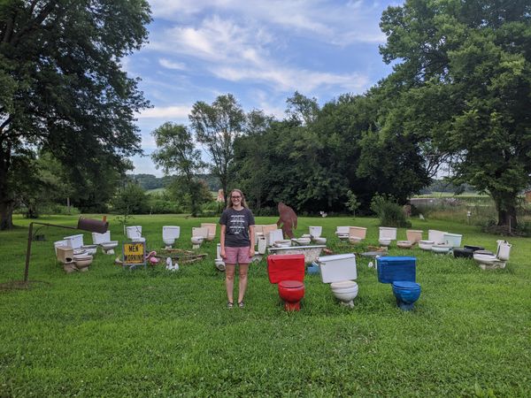 Cassey, with long blonde hair, stands in a grassy lot next to 3 toilets painted red, white, and blue respectively. In the background is a semi-circle of more old toilets, a mailbox, a sign that says 'MEN WORKING', and a life-size rusty metal yeti figure.