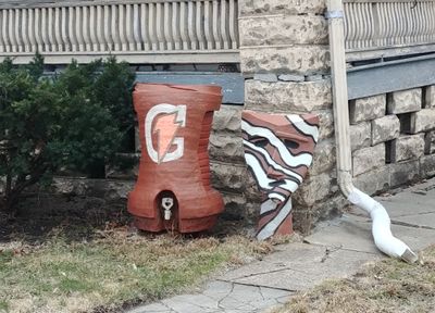 on the ground at the corner of a porch sits two clay sculptures, each about 2-3 feet tall. one is of a team-sized gatorade dispenser complete with painted logo, and the other is of a triangle shaped mask and covered in painted zebra stripes