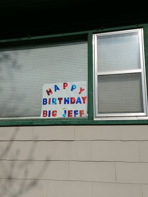 a sign with multicolored letters hangs in the window of a building. the sign reads 'Happy birthday big Jeff' in all caps