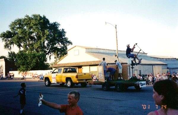 a bright yellow truck pulls a pretty bare-bones trailer that has a slingshot contraption mounted on the back, with a guy riding the contraption with feet on the pedals, about to launch a hot dog