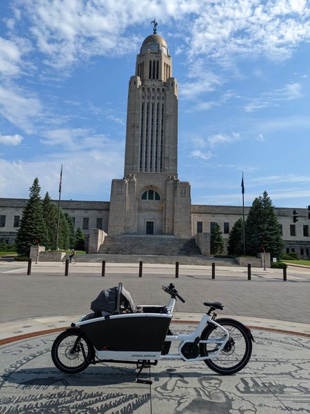 An Urban Arrow bike, a bakfiet-style cargo bike with a white frame and a big black bucket on the front, sits in front of the Nebraska State Capitol building. There's an infant car seat sticking out of the bucket